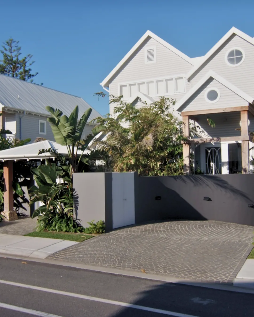 Street view of a grey weatherboard house with a dark rendered wall and cobblestone driveway
