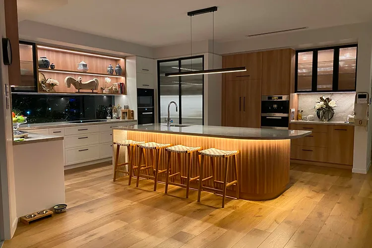 Kitchen with a curved timber island with warm under-lighting, timber stools and oak floors