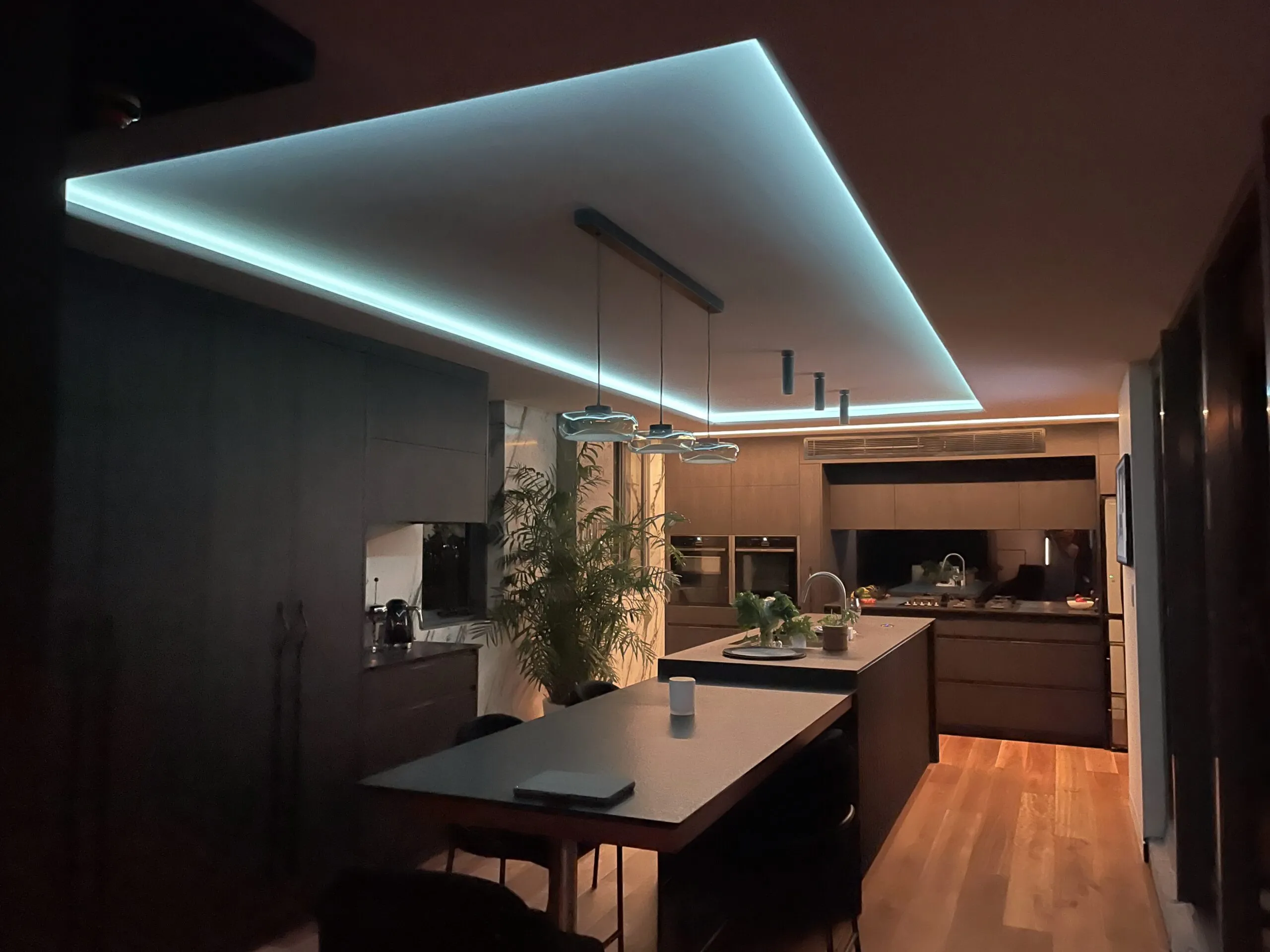 Dark kitchen with a glowing blue ceiling light line over the island and warm timber floors