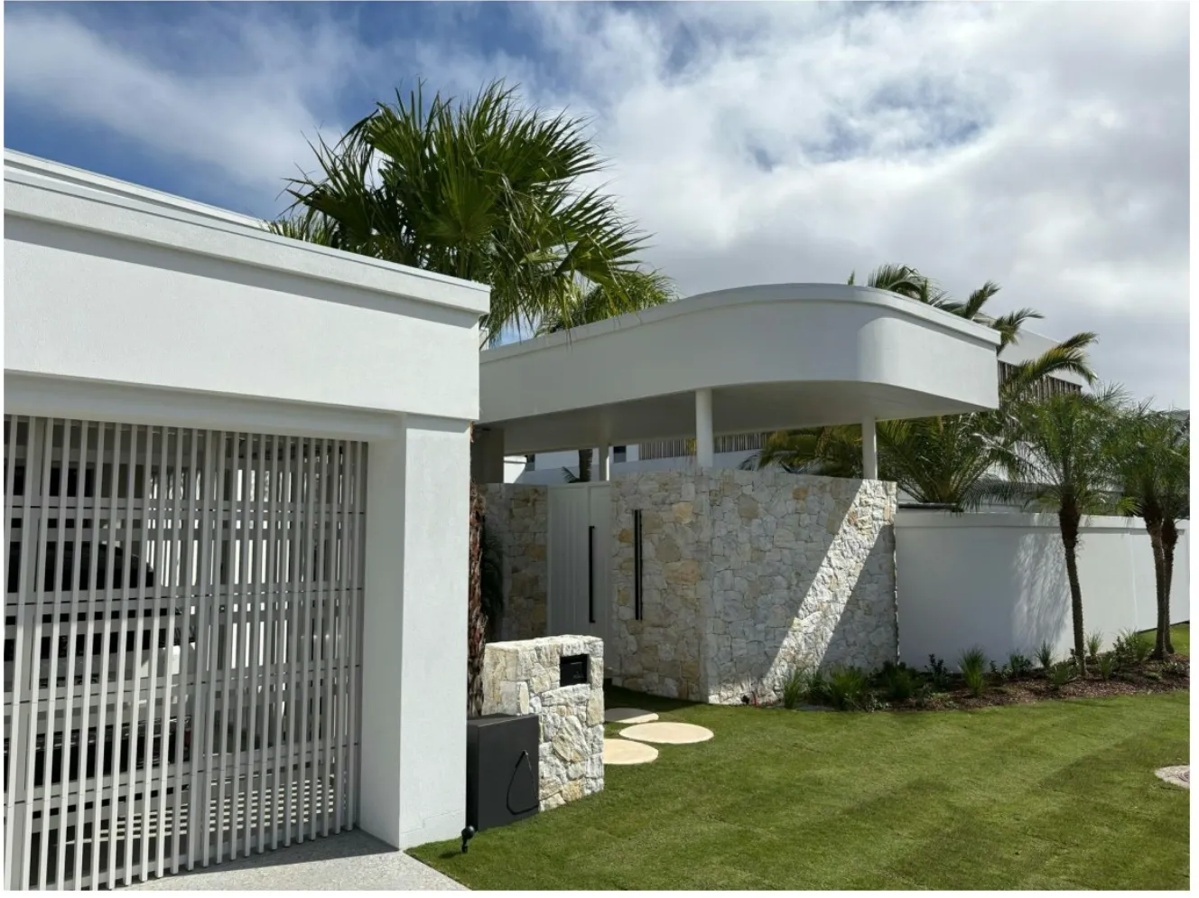 Modern white home with a slatted gate, curved roofline and stone wall beside a lawn