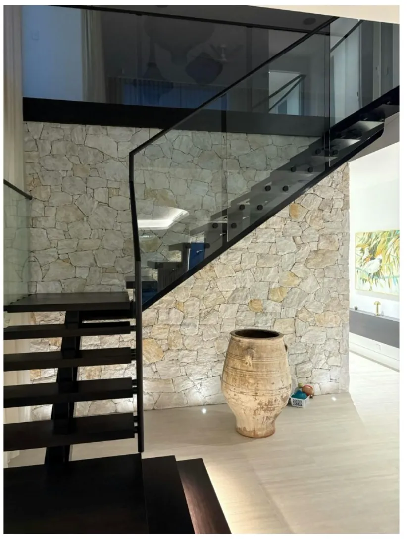 Floating black staircase with a glass balustrade beside a stone feature wall and a large urn