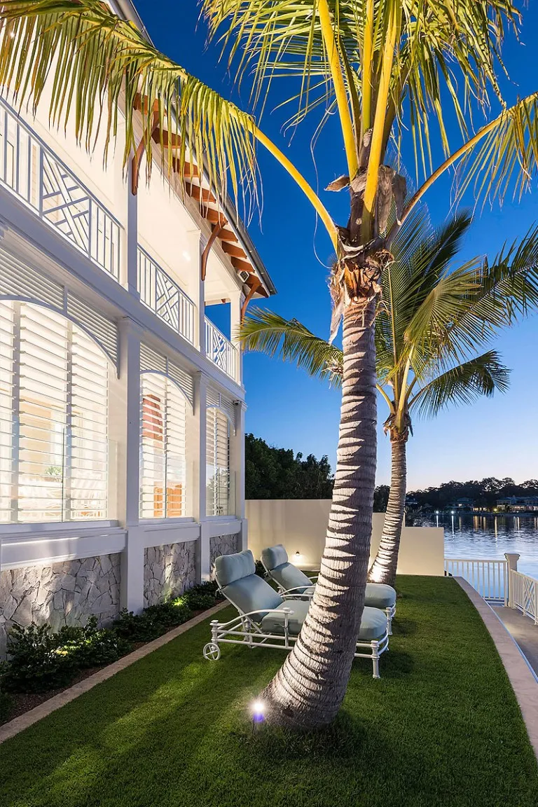 Waterfront home with white shutters and tall palm trees on the lawn at dusk